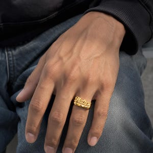 May include: A gold-colored ring with the name "Matthew" engraved in a cursive font. The ring is worn on a finger, and the background includes a person wearing a black top and blue jeans.
