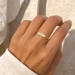 Op de afbeelding: Een goudkleurige ring met een rechthoekig oppervlak, gegraveerd met de tekst "VedawoodNYC". De ring wordt aan een vinger gedragen en toont zijn eenvoudige ontwerp. Het gepolijste oppervlak van de ring reflecteert licht, wat de gladde textuur benadrukt.