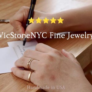 May include: A person writing on a white card with a black pen, wearing two gold rings. The image includes the text "VicStoneNYC Fine Jewelry" and "Handmade in USA". Five gold stars are visible.