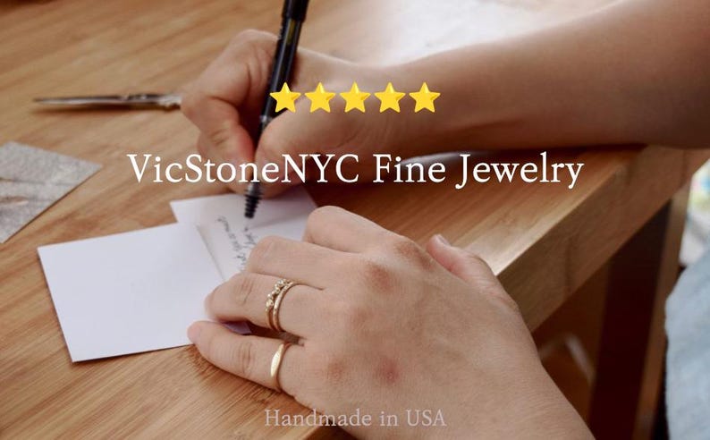 May include: A person writing on a white card with a black pen, wearing two gold rings. The image includes the text "VicStoneNYC Fine Jewelry" and "Handmade in USA". Five gold stars are visible.