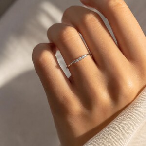 May include: A delicate gold ring featuring three small, sparkling diamonds set in a row. The ring is thin and elegant, designed for everyday wear. The ring is shown on a person's finger.