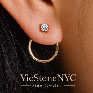 Puede incluir: Primer plano de una oreja adornada con dos joyas de oro. Un pequeño pendiente de botón redondo y transparente está en la parte superior de la oreja, y un pendiente de aro dorado está debajo. El texto "VicStoneNYC Fine Jewelry" está en la parte inferior.