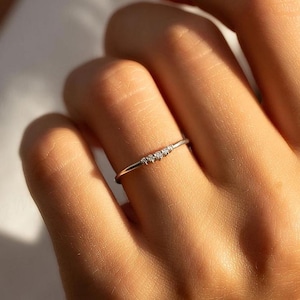 May include: A delicate silver ring with three small, clear gemstones set in a row. The ring is thin and minimalist, designed to be worn on a finger. The ring is shown on a person's finger.