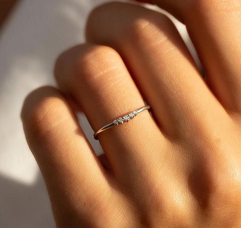 May include: A delicate silver ring with three small, clear gemstones set in a row. The ring is thin and minimalist, designed to be worn on a finger. The ring is shown on a person's finger.