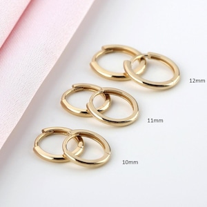 May include: Three pairs of gold-colored hoop earrings in varying sizes: 10mm, 11mm, and 12mm. The earrings are displayed on a white surface with a pink fabric in the background. The hoops have a simple, classic design.