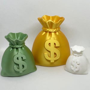 May include: Three money bag sculptures in green, gold, and white. Each bag has a dollar sign on the front. The bags vary in size, with the largest in gold and the smallest in white.