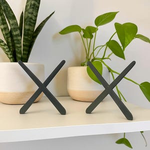 K&ouml;nnte beinhalten: Zwei dunkelgraue dekorative Holzobjekte in X-Form. Die Objekte befinden sich auf einem wei&szlig;en Regal, im Hintergrund befinden sich Topfpflanzen. Die Pflanzen haben gr&uuml;ne Bl&auml;tter und stehen in wei&szlig;en T&ouml;pfen mit hellbraunem Sockel.