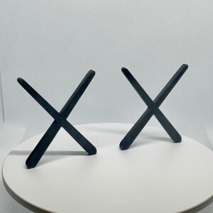 K&ouml;nnte beinhalten: Zwei schwarze, X-f&ouml;rmige Dekorationsgegenst&auml;nde. Jeder Gegenstand besteht aus einem festen Material und hat eine matte Oberfl&auml;che. Die Objekte sind auf einer wei&szlig;en Oberfl&auml;che vor einem wei&szlig;en Hintergrund positioniert.