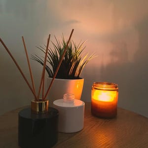May include: Three decorative containers on a wooden table. A black diffuser with gold accents and wooden sticks, a white diffuser, and a glass jar with a lit candle inside.