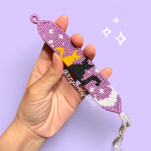 May include: A handmade friendship bracelet in shades of purple, white, black, and yellow. The bracelet features a witch design with stars and a crescent moon. The text "@itzylovelyshop" is visible on the bracelet.