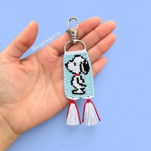 May include: A light blue Snoopy keychain with a cartoon image of Snoopy. The keychain has white tassels with red accents and a silver clasp. The keychain is held in a hand against a blue background.