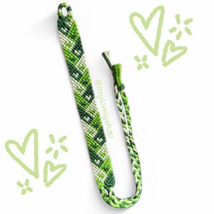 May include: Green and white friendship bracelet with a chevron pattern. The bracelet is tied with a knot at the end. The text "@izzy.lovely.shop" is written on the bracelet.