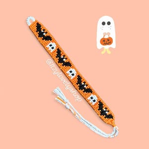 May include: An orange and black Halloween friendship bracelet with a pattern of bats and skulls. The bracelet has a white tassel and the text "@itzylovelyshop". A cartoon ghost holding a pumpkin is also in the image.