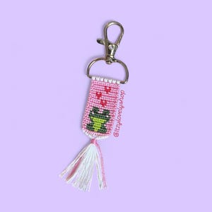 May include: A pink handmade keychain with a frog design. The keychain features a green and black frog, red hearts, and a white and pink tassel. It is attached to a silver-colored clasp.