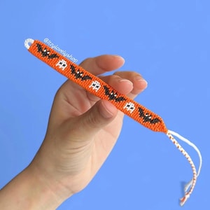 May include: An orange friendship bracelet with a repeating pattern of black bats and white ghosts. The bracelet is held against a blue background. The text "@itzylovely shop" is visible.