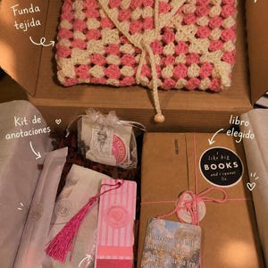 Puede incluir: Una caja de regalo con una funda de libro tejida en rosa y crema, un libro envuelto con una pegatina I like big BOOKS and i cannot lie, un marcapáginas de rayas rosas con borla y una bolsa pequeña de artículos de papetería con etiquetas.