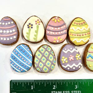 May include: Eight Easter egg-shaped cookies decorated with pastel colors and various patterns, including stripes, dots, and flowers. The cookies are arranged on a white surface with a ruler for scale.