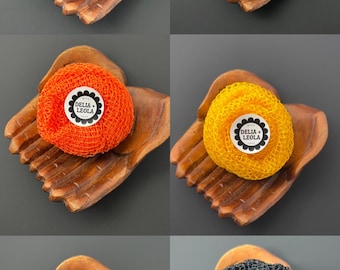 Op de afbeelding: Een collectie van zes ronde gaassponzen in blauw, groen, oranje, geel, rood en zwart. Elke spons heeft in het midden een wit rond label met de tekst DELIA + LEOLA. De sponzen rusten op individuele houten schaaltjes.