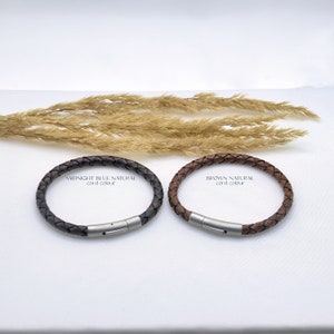 May include: Two leather bracelets with a braided design. The bracelet on the left is dark blue and the bracelet on the right is brown. Both bracelets have a silver clasp.