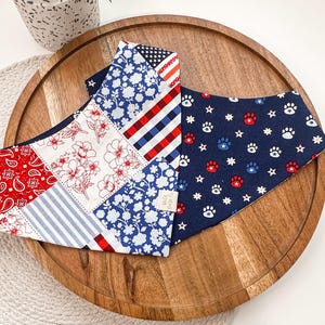 May include: Two dog bandanas with patriotic patterns. One features a patchwork design with red paisley, white floral, blue floral, and striped patterns. The other is navy blue with paw prints and stars in red, white, and blue.