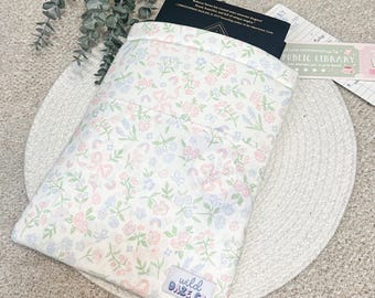 May include: A floral print book sleeve with a pastel color scheme. The sleeve features a pattern of pink bows, blue flowers, and green leaves on a white background. A book is partially visible inside the sleeve. The sleeve has a tag that says "wild daze co."