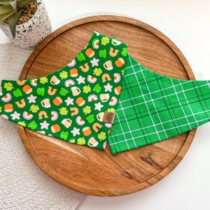 May include: Two green fabric dog bandanas. One bandana has a green, white, and orange pattern with shamrocks, stars, rainbows, and beer mugs. The other bandana has a green and white plaid pattern. Both bandanas have a brown label that says "Izzy & Paco".