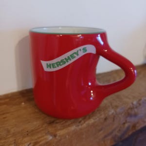 May include: A red ceramic mug with a curved handle and the Hershey's logo in white. The mug has a green rim and sits on a wooden surface. The mug is a vibrant red color.