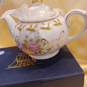 Può includere: Una teiera in porcellana bianca con un motivo floreale di rose rosa, bordi dorati e un pomello dorato sul coperchio. La teiera poggia su una scatola blu scuro con la scritta dorata Nantucket e il disegno di un veliero stampato sul coperchio.