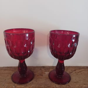 Peut inclure: Deux verres à pied en verre rouge profond assortis, présentant un motif en nid d'abeille texturé sur le calice. Chaque verre possède une tige lisse et arrondie ainsi qu'une base circulaire, fabriqués en verre rubis translucide.