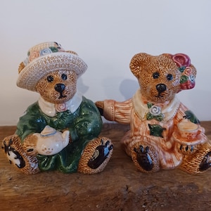 Puede incluir: Dos figuras de osos de peluche de cerámica. Un oso viste un vestido verde y un sombrero, sosteniendo una tetera. El otro oso viste un vestido naranja y sostiene una taza de té. Ambos osos tienen ojos negros y pies en forma de pata.