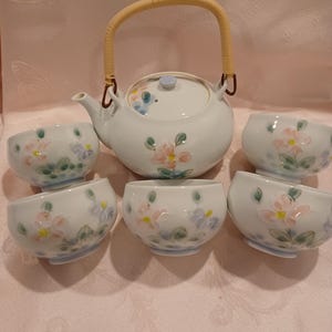 May include: A ceramic tea set consisting of a teapot with a light-colored woven handle and four matching tea cups. Each piece is white with a hand-painted floral design featuring soft pink and blue petals with green leaves on the exterior.
