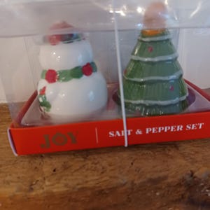 May include: A Christmas-themed salt and pepper set. One shaker is a snowman with a red hat and green scarf, the other is a green Christmas tree. The set is in a clear plastic box with a red base that reads "JOY | SALT & PEPPER SET."