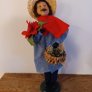 May include: A decorative figurine of a man wearing a straw hat, red cape, and denim shirt. The figurine holds a red flower and a woven basket with greenery. The figure stands on a green base and has black shoes.
