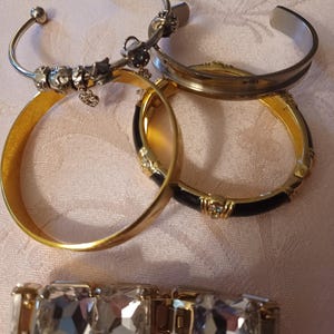 Pode incluir: Uma coleção de quatro pulseiras, incluindo um bracelete prateado com berloques, um bracelete prateado liso, um bracelete dourado com esmalte preto e pedras transparentes, e uma pulseira de elos dourados com três grandes cristais quadrados transparentes.