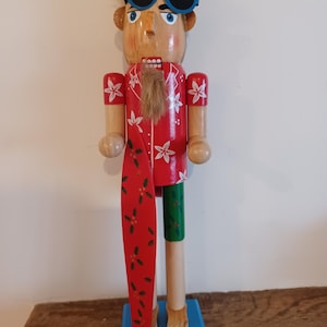May include: Wooden nutcracker figurine of a surfer wearing blue sunglasses and a red Hawaiian shirt with white flower designs. The figurine holds a red surfboard with green leaves and has one green leg. It stands on a blue base.