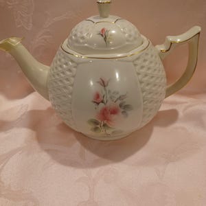 May include: A cream-colored porcelain teapot featuring a raised diamond-textured pattern on the body and lid. The teapot is decorated with a painted pink rose and green leaves, accented with gold trim on the handle, spout, lid, and rim.