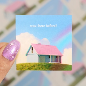 May include: A square sticker featuring a small house with a pink roof and a blue exterior, set against a sky with a rainbow and clouds. The text "was i here before?" is above the house. The sticker is held by a hand with a sparkly purple nail.