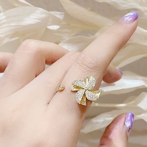 May include: A gold ring with a flower design featuring clear rhinestones. The ring is open on one side and has a small, round rhinestone on the other side.