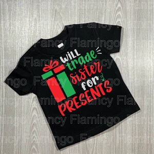 Peut inclure: T-shirt noir avec un graphique de boîte-cadeau rouge et verte et le texte "Will trade sister for presents".