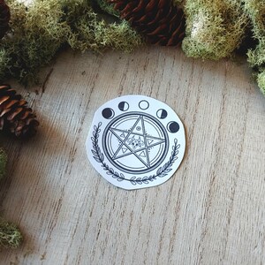 May include: A white sticker with a black pentagon star design, surrounded by five crescent moons and a leafy border. The star has a smaller pentagon inside with a circle in the center.