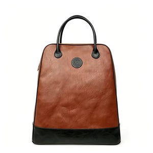 May include: A brown backpack with a smooth, textured finish and a solid black base. The bag features two black top handles, black piping along the edges, and a circular black patch on the front centre with the letters AK embossed in silver.
