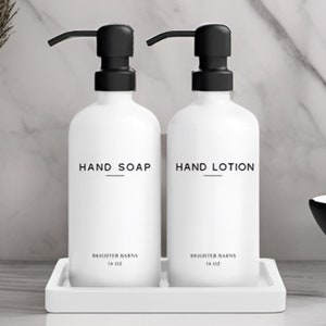 May include: Two white pump bottles with black pumps and labels that read "Hand Soap" and "Hand Lotion". The bottles are on a white tray and are in front of a white marble background.