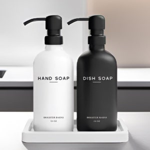 May include: Two black and white pump bottles with labels that read "Hand Soap" and "Dish Soap". The bottles are on a white tray and are sitting on a white countertop.