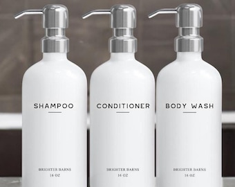 May include: Three white pump bottles labeled "SHAMPOO", "CONDITIONER", and "BODY WASH" with silver pump tops. Each bottle has the text "BRIGHTER BARNS" and "16 OZ" printed on the front. The bottles are arranged on a gray surface.