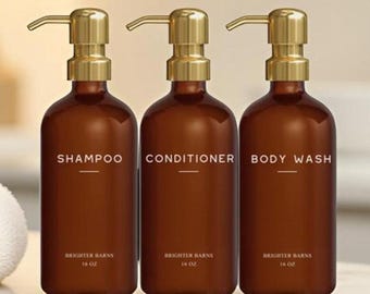 May include: Three brown pump bottles labeled "SHAMPOO", "CONDITIONER", and "BODY WASH" with gold-colored pumps. The bottles are on a white surface with a white towel to the left. The text "BRIGHTER BARNS" and "16 oz" are below the labels.