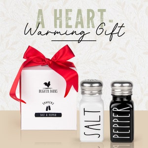 Puede incluir: Una caja de regalo blanca con un lazo rojo y un logotipo de gallo. La caja dice "Brighter Barns" y "Saleros y pimenteros". Dos saleros y pimenteros de cer&aacute;mica, uno blanco y uno negro, con las palabras "Sal" y "Pimienta" escritas en ellos.