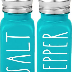 Puede incluir: Un juego de dos saleros y pimenteros de vidrio azul turquesa con tapas plateadas. Los saleros tienen letras blancas que dicen "SALT" y "PEPPER".