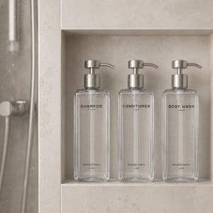 May include: Three clear glass bottles with silver pump tops labeled "SHAMPOO", "CONDITIONER", and "BODY WASH". Each bottle has a capacity of 16 oz. The bottles are displayed in a recessed shelf in a neutral-toned bathroom.