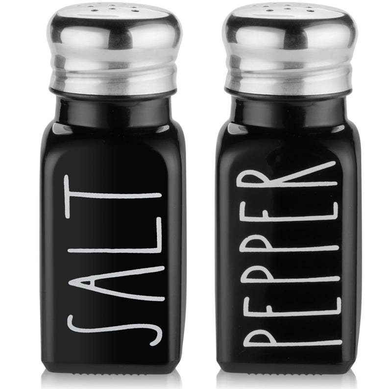 Puede incluir: Salero y pimentero de vidrio negro con tapas plateadas. Los shakers tienen letras blancas que dicen "SALT" y "PEPPER".