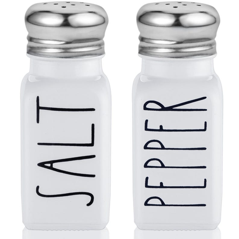 Puede incluir: Salero y pimentero de cer&aacute;mica blanca con tapas plateadas. Los saleros tienen letras negras que dicen "SALT" y "PEPPER".